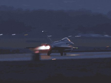 a fighter jet is flying over a field with a sunset in the background