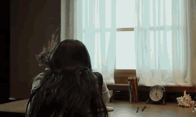 a woman is sitting at a desk in front of a window with a clock on it .