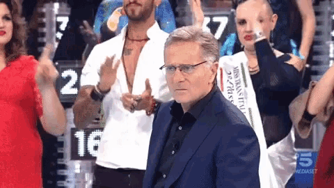 a man in a suit stands in front of a group of people including a woman wearing a miss italy crown