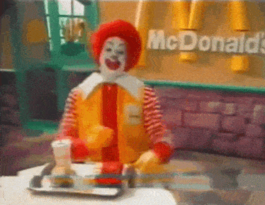 a mcdonald 's clown is sitting at a table holding a tray of food .