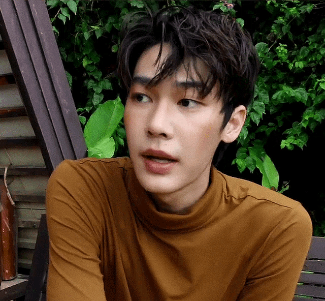 a young man wearing a brown turtleneck is sitting on a wooden bench