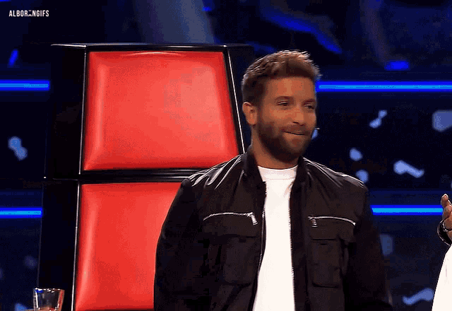 a man in a black jacket sits in front of a red chair with the gifs written above him