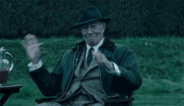 a man in a suit and hat is sitting in a chair waving .