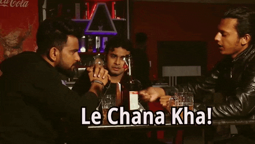 three men are sitting at a table and one of them is holding a bottle of beer and says " le chana kha "