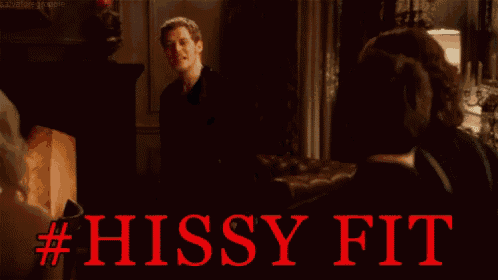 a man in a black shirt is talking to another man in a dark room with the words #hissy fit written in red