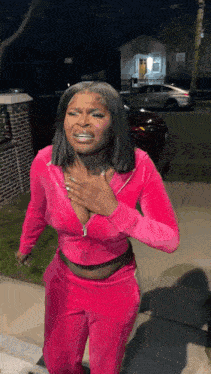 a woman in a pink tracksuit is standing on a sidewalk at night with her hand on her chest .