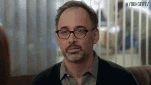 a man with glasses and a beard is sitting on a couch in front of a younger tv sign .