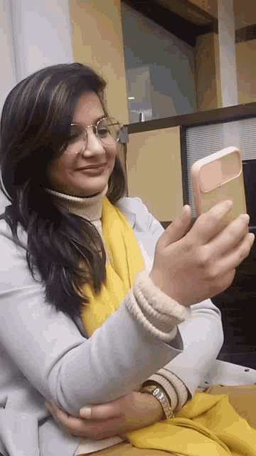 a woman wearing glasses and a yellow scarf takes a selfie with her phone