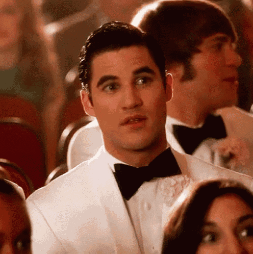 a man in a white suit and black bow tie is sitting in a crowd of people