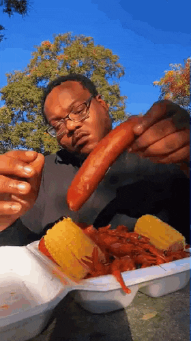 a man with glasses is eating a hot dog and corn on the cob .