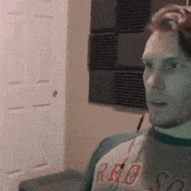 a man wearing a red sox shirt is sitting in a room with a door in the background .