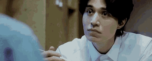 a man in a white shirt is eating noodles with chopsticks and looking at another man .