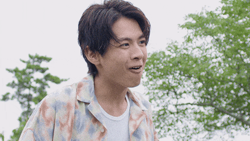 a young man wearing a colorful shirt and a white shirt stands in front of trees