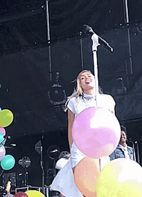 a woman in a white dress is singing into a microphone surrounded by balloons