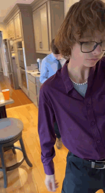 a young man wearing glasses and a pearl necklace is standing in a kitchen