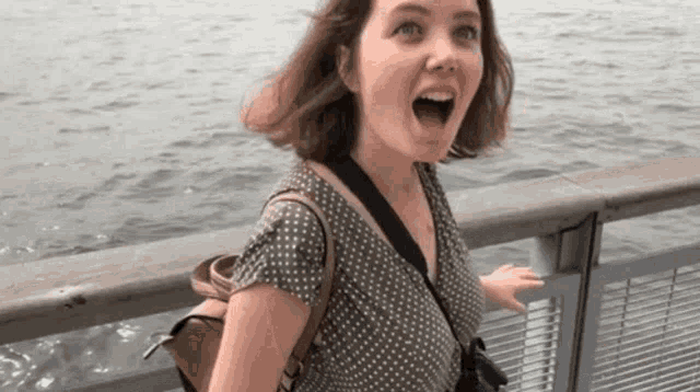 a woman with a backpack is standing on a bridge overlooking a body of water with her mouth open .