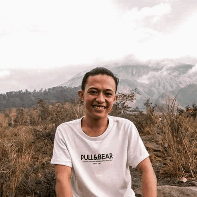 a young man wearing a white pull & bear t-shirt is sitting on a rock in a field .