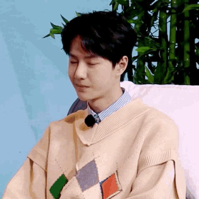 a young man wearing a sweater with a microphone on his neck is sitting on a bed with his eyes closed