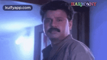 a man with a mustache is standing in front of a door and looking at the camera .