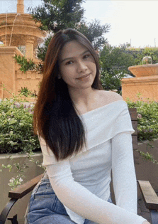 a woman in a white off the shoulder top sits on a bench in front of a fountain