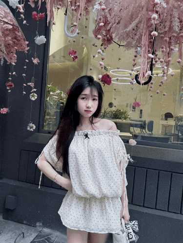 a woman wearing a white off the shoulder top stands in front of a window with pink flowers hanging from it
