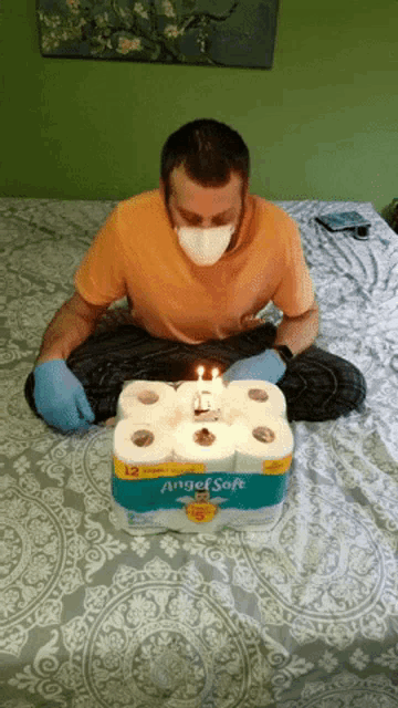 a man wearing a mask is sitting on a bed next to a box of angelsoft toilet paper