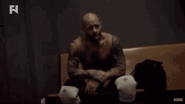 a man without a shirt is sitting on a bench in a locker room with boxing gloves .