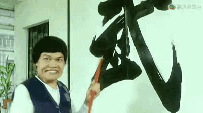 a man is smiling while holding a brush in front of a wall with chinese writing on it