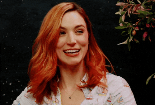 a woman with red hair is smiling and wearing a shirt that has sailboats on it