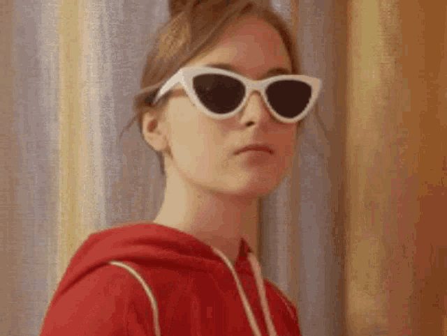 a woman wearing a red hoodie and white sunglasses is looking at the camera .
