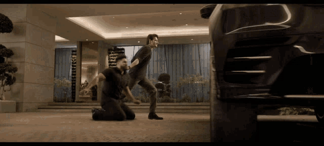 two men kneeling in front of a black car in a lobby