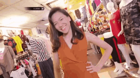 a woman in an orange dress is standing in a clothing store with her hands on her hips .