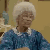 an elderly woman in a blue dress and glasses is sitting at a table .