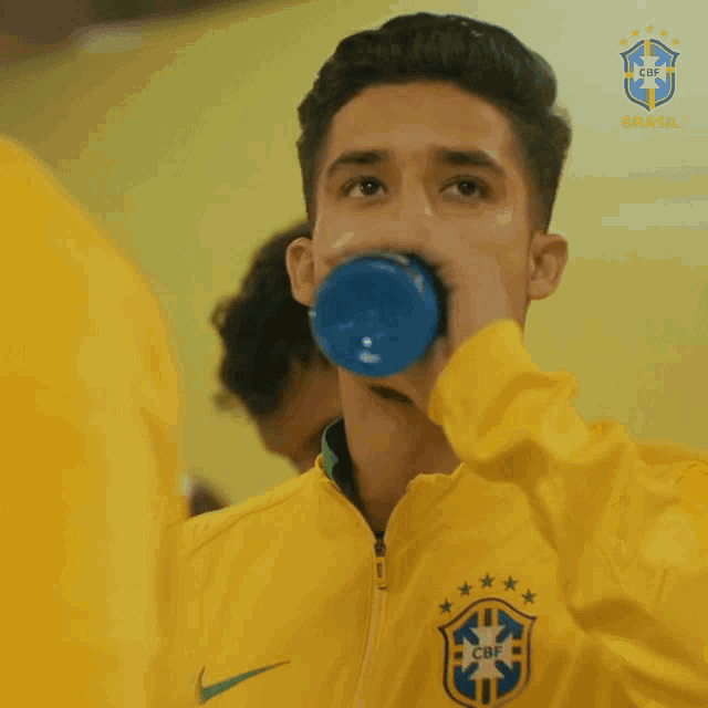a young man in a yellow jacket drinking from a blue bottle with the cbf logo behind him