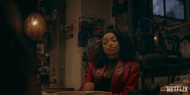 a woman in a red jacket sits at a desk with a netflix logo on the bottom right