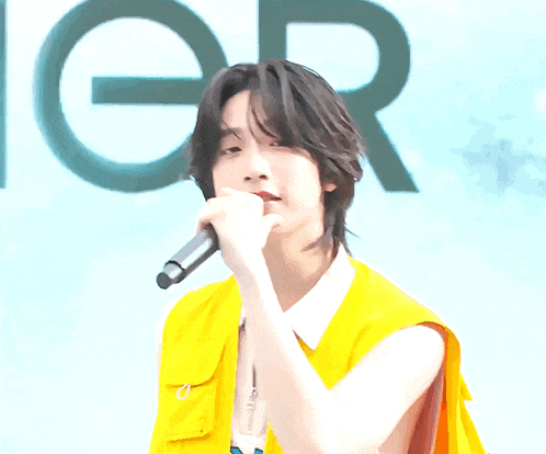 a man in a yellow vest sings into a microphone in front of a sign that says n.e.i.