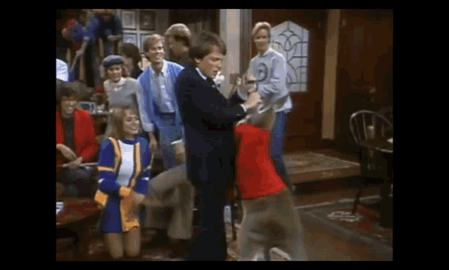 a group of people are dancing in a living room while a man in a suit holds a dog .
