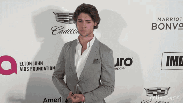 a man in a suit stands in front of an elton john aids foundation advertisement