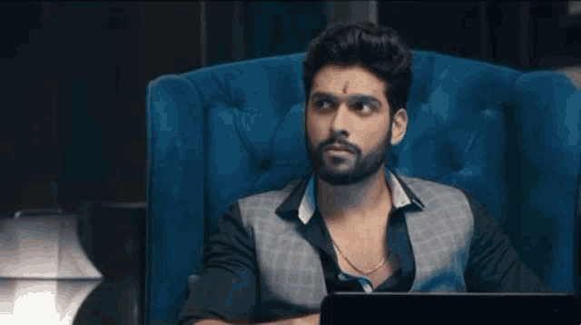 a man with a beard is sitting in a blue chair looking at a laptop .