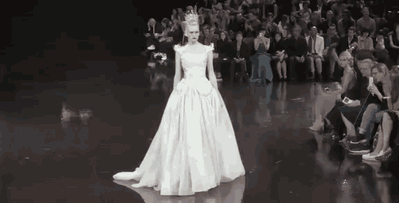 a woman in a white dress with a crown on her head is walking down a runway .