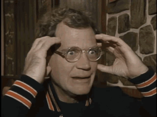 a man wearing glasses and a striped sweater holds his hands to his head