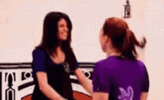 two women are shaking hands in front of a white wall . one of the women is wearing a purple shirt .