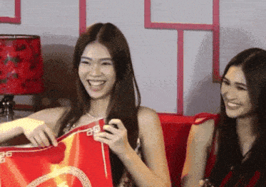 two women are sitting on a red couch and one of them is holding a red bag with a greek key pattern on it