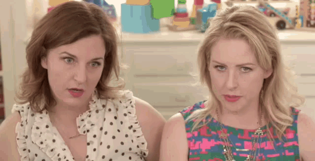 two women are sitting next to each other and making funny faces . one of the women is wearing a polka dot shirt .