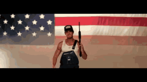 a man in overalls is holding a gun in front of a large american flag