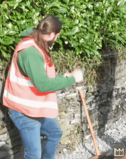 a man wearing an orange vest and a green sweater is sweeping a stone wall