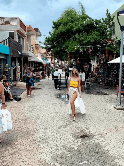 a woman in a yellow top and white skirt walks down a busy street with a bag that says ' i love you '
