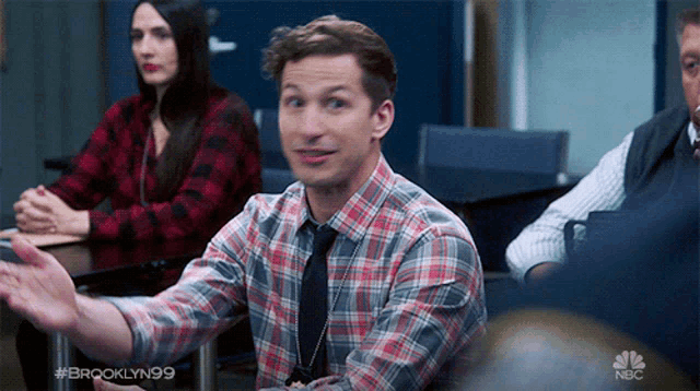 a man in a plaid shirt and tie is sitting in a classroom with brooklyn99 in the corner
