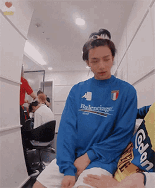 a man wearing a blue balenciaga sweatshirt sits in a room with other people