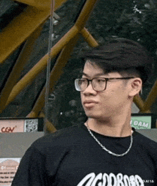 a young man wearing glasses and a necklace is standing in front of a cgv building .